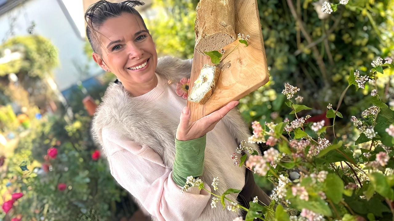 Kräuterhexe Uschi Zezelitsch hält ein Holzbrett mit Käse und Brot aus Buchweizen