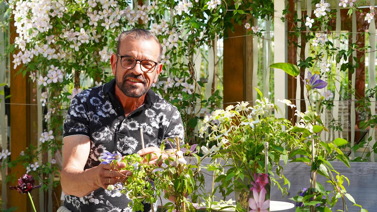 Karl Ploberger sitzt in einem Garten vor einer Clematis-Pflanze mit weißen Blüten.