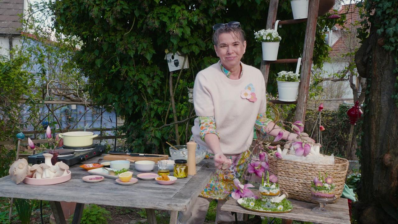 © ORF/Uschi Zezelitsch Uschi Zezelitsch steht an einem Holztisch im Garten und präsentiert ihre „Frühlings-Glückspilze“ im Glas“ als Antipasti.