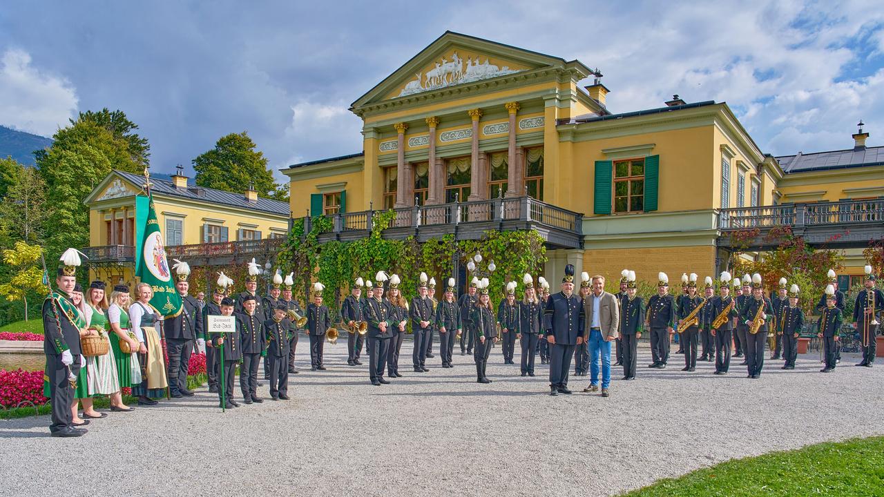Hans Knauß und die Salinenmusikkapelle vor der Kaiservilla in Bad Ischl. Daneben stehen Frauen in Dirndln und ein Bannerträger.