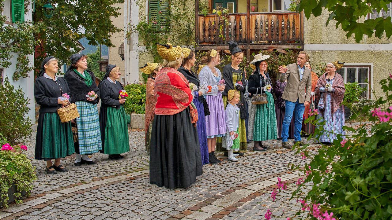 Die Goldhauben- und Kopftuchgruppe in farbenfroher, traditioneller Festtagstracht auf einem gepflasterten Platz zwischen historischen Häusern unterhalten sich mit Hans Knauß.