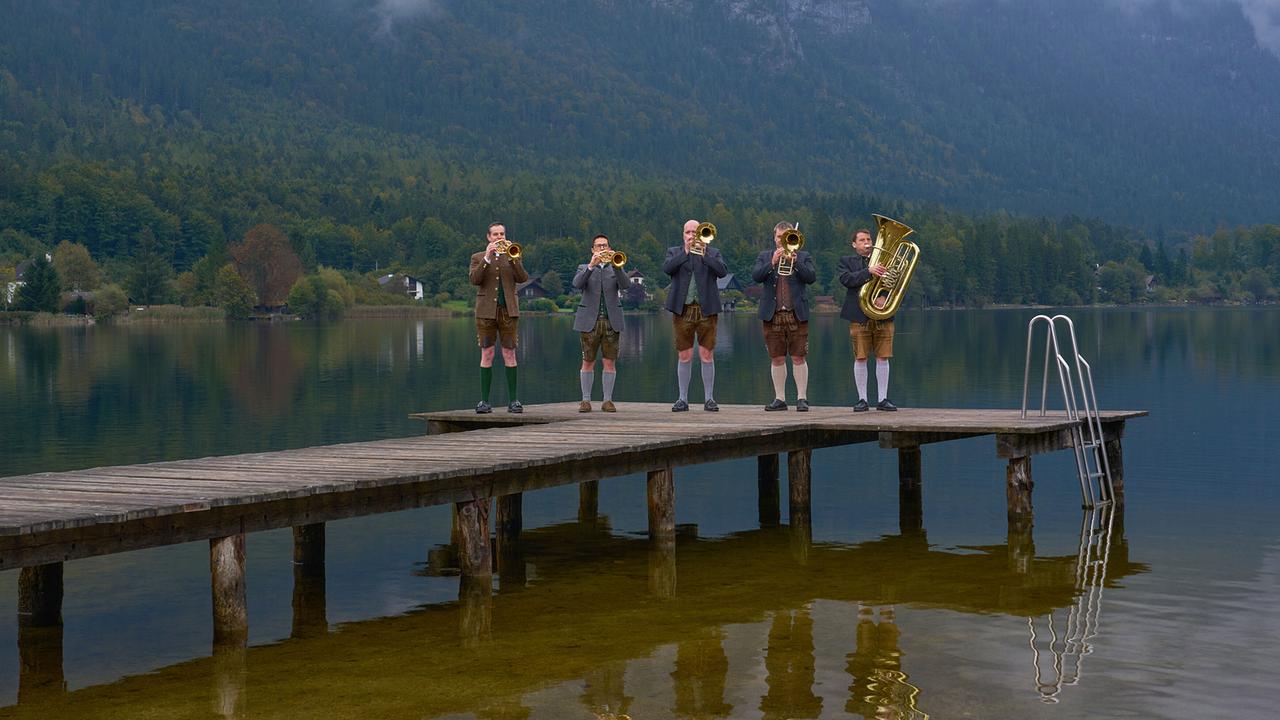 Die Steff’n Hansl Musi in Tracht spielen Trompeten, Flügelhorn und Tuba auf einem Holzsteg über einem klaren See, umgeben von Bergen.