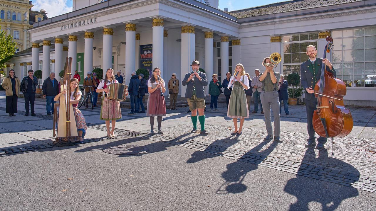 Die SOS Musi spielt Harfe, Akkordeon, Blockflöten, Trompete und Kontrabass vor einem klassizistischen Gebäude einer Kurstadt, während Passanten zuschauen.