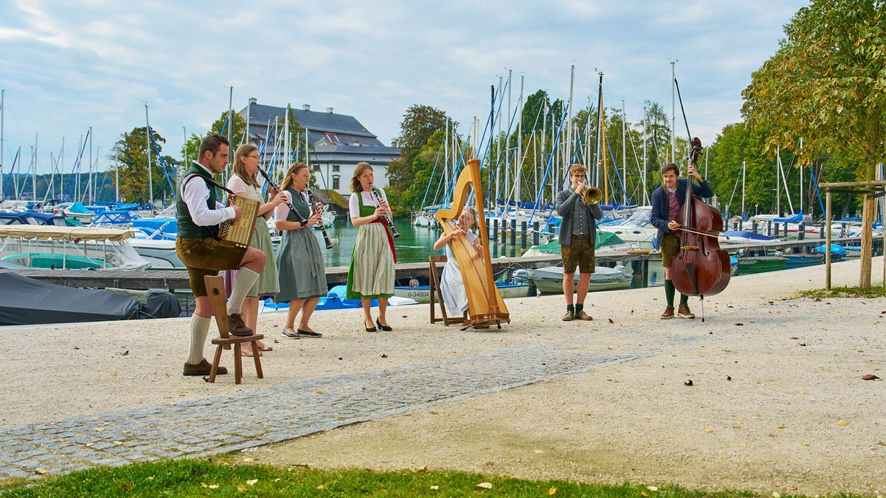 Die BruckZuck Musi in Tracht spielt Instrumente wie Akkordeon, Klarinette, Harfe, Trompete und Kontrabass am Ufer eines Hafens mit vielen Segelbooten.