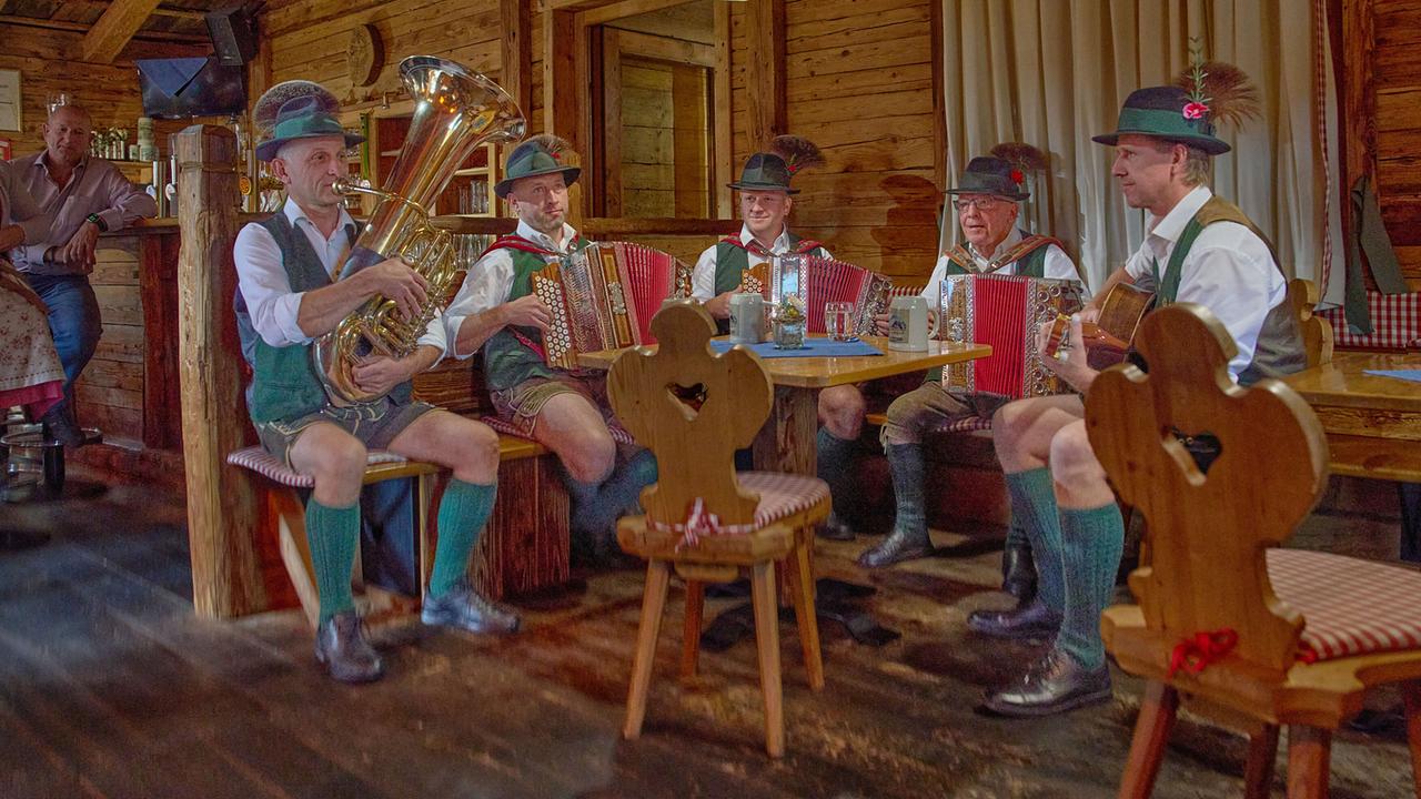 Die Aberseea Musikanten in Lederhosen und grünen Westen spielen Akkordeons, Tuba und Gitarre in einer rustikalen Stube mit Holzvertäfelung.