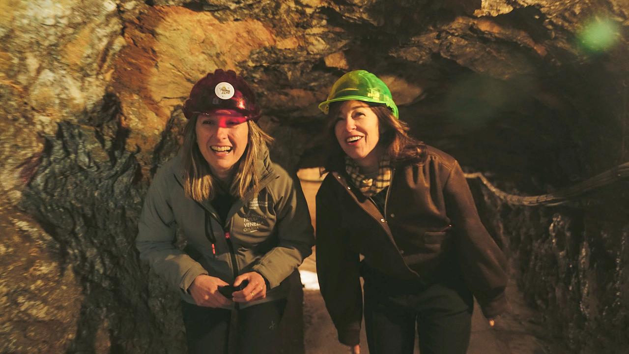 Julia Cencig im Gespräch mit Bergwerksführerin Leila Meroi, beide mit Schutzhelmen, in einem niedrigen Stollengang im Bergwerk Cave del Prebil