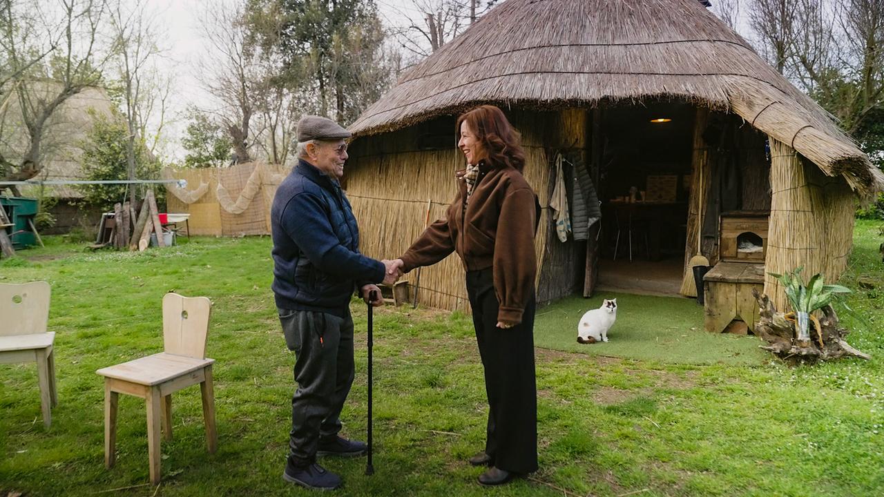 Julia Cencig begrüßt Luigino Marchesan, ein Fischer aus Caorle, in einem Garten vor einer kleinen, strohgedeckten Hütte.