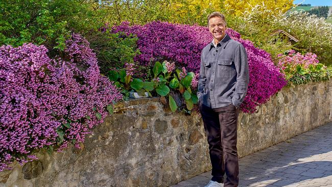 Hans Knauß steht auf einem Gehweg neben einer Steinmauer mit üppig blühenden Sträuchern bei Sonnenschein.