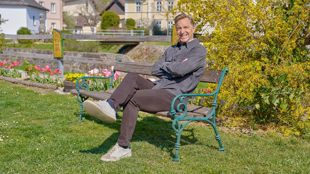 © ORF/Alois Kuschetz Hans Knauß sitzt mit verschränkten Armen auf einer Parkbank in einem gepflegten Garten bei Sonnenschein.