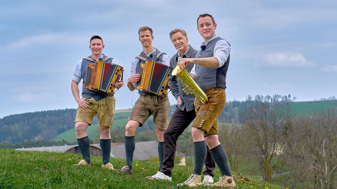 © ORF/Alois Kuschetz Die drei Musiker der Gruppe ZamQuetscht mit Ziehharmonikas und einem Blasinstrument stehen mit Hans Knauß auf einer Wiese.