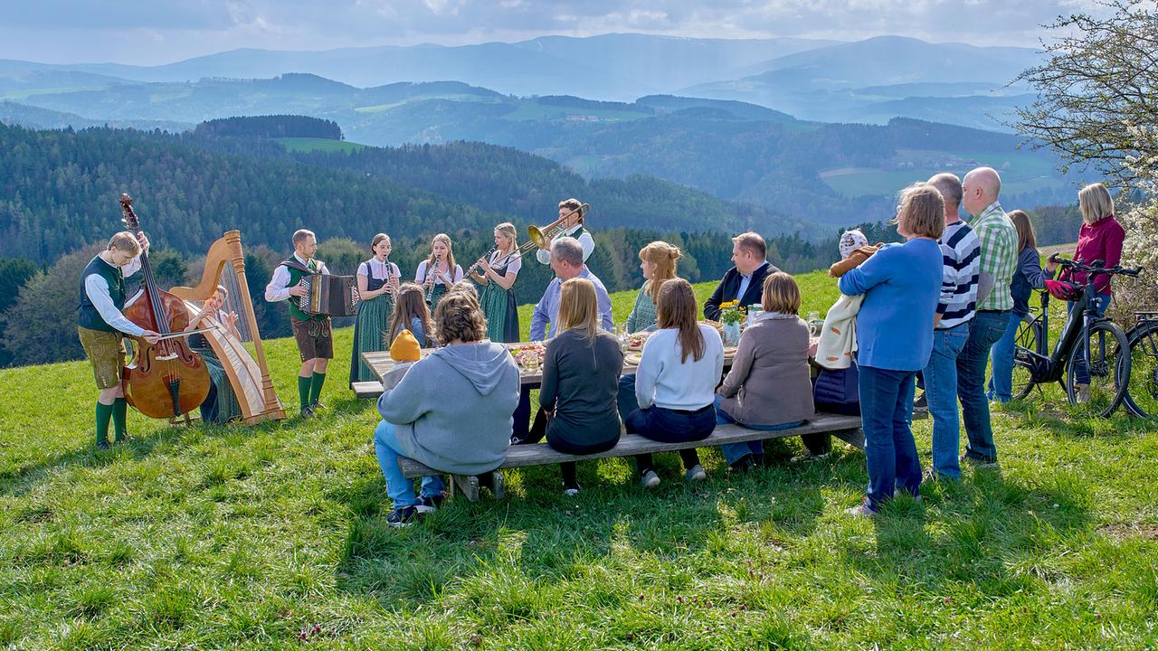 © ORF/Alois Kuschetz Die Gruppe Vollholzmusi spielen im Freien, während eine Gruppe von Menschen an einem Holztisch die Darbietung in der Natur verfolgt.