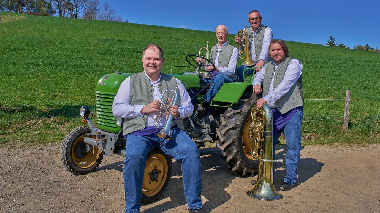 © ORF/Alois Kuschetz Quadra Buckliga, vier Männer in Tracht, sitzen und stehen mit Blasinstrumenten auf und neben einem grünen Traktor auf einem Feldweg.