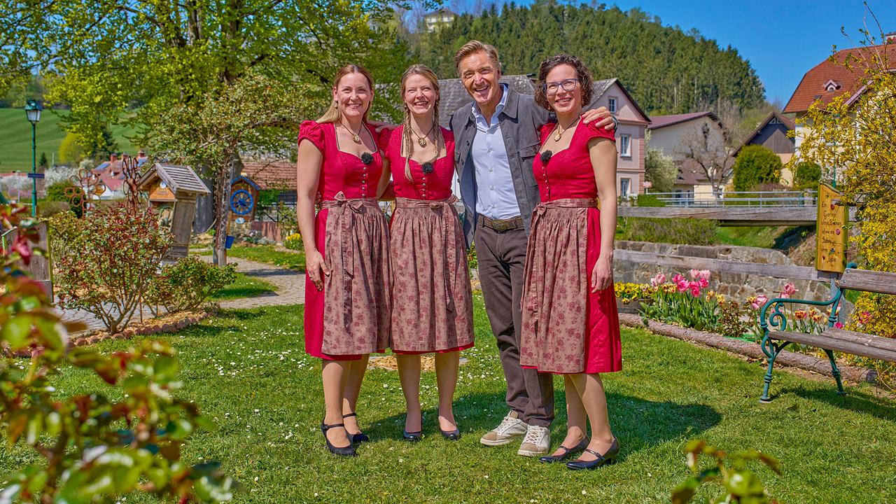 © ORF/Alois Kuschetz KAKtussis, drei Frauen in Dirndln, und Hans Knauß stehen eng in einem sonnigen Garten vor ländlicher Umgebung.