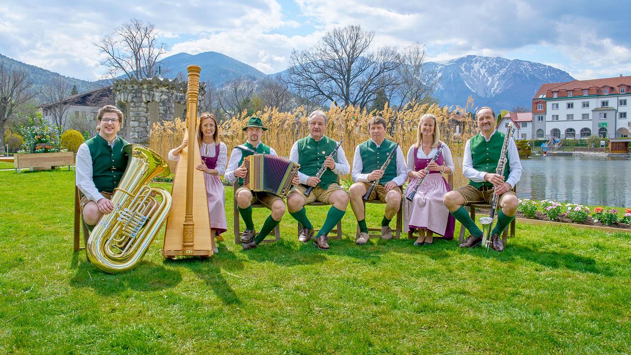 © ORF/Alois Kuschetz Die Frohnberger Klarinettenmusi, eine Volksmusikgruppe in Tracht, posiert mit Blas- und Saiteninstrumenten auf einer grünen Wiese vor Bergkulisse.