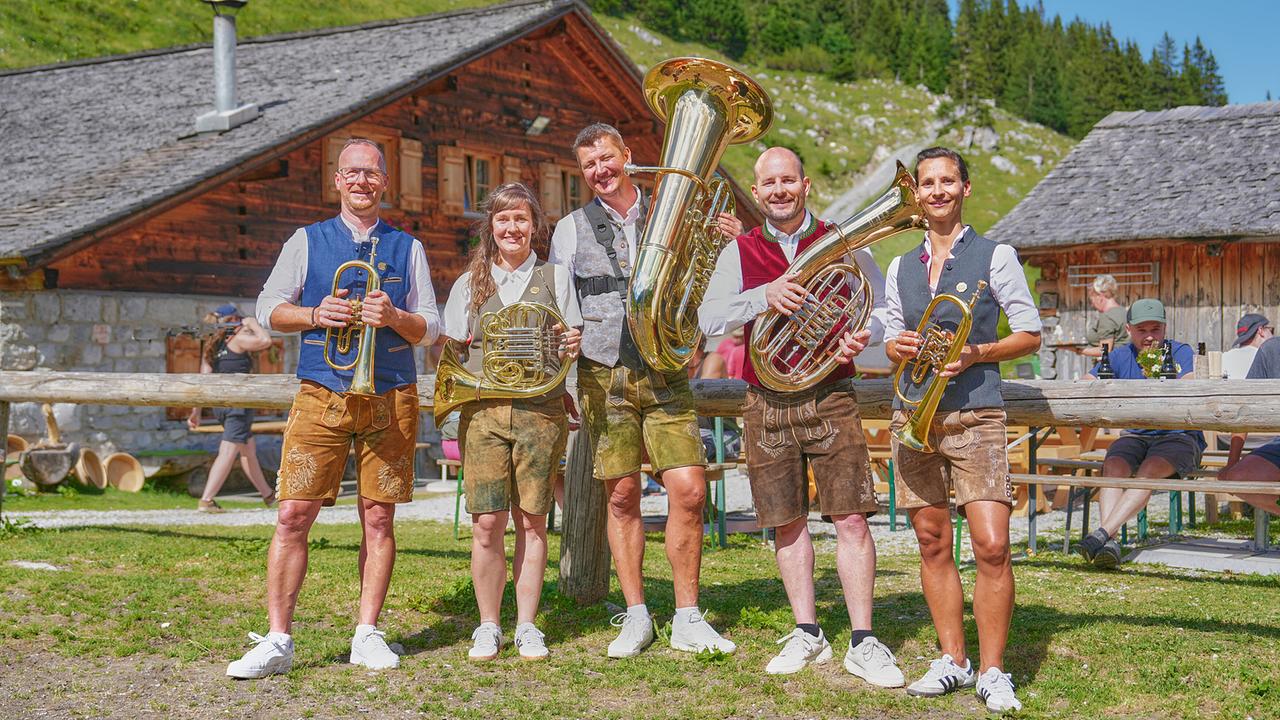 Die "5 Kant Musik", fünf Musiker:innen in Tracht, präsentieren vor einer Almhütte ihre Blasinstrumente.