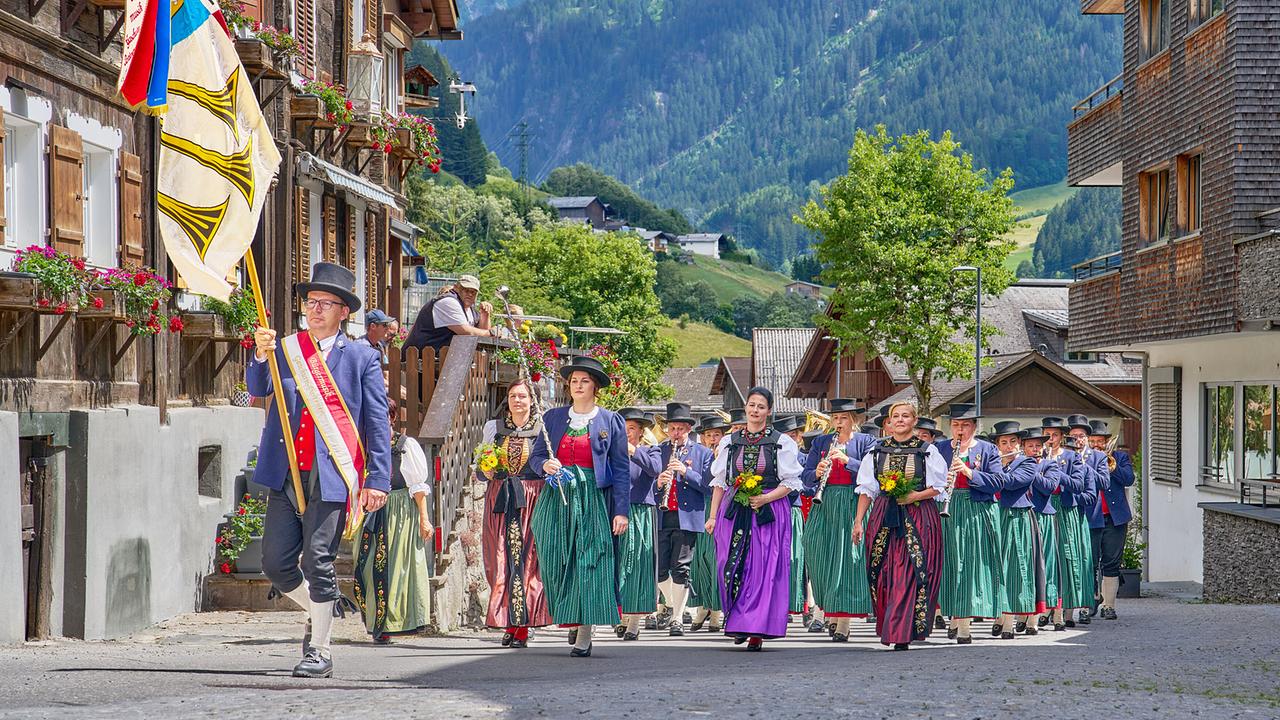 Die Bürgermusik Gaschurn-Partenen in Tracht zieht mit Musikkapelle bei einem Festzug durch ein Bergdorf. 