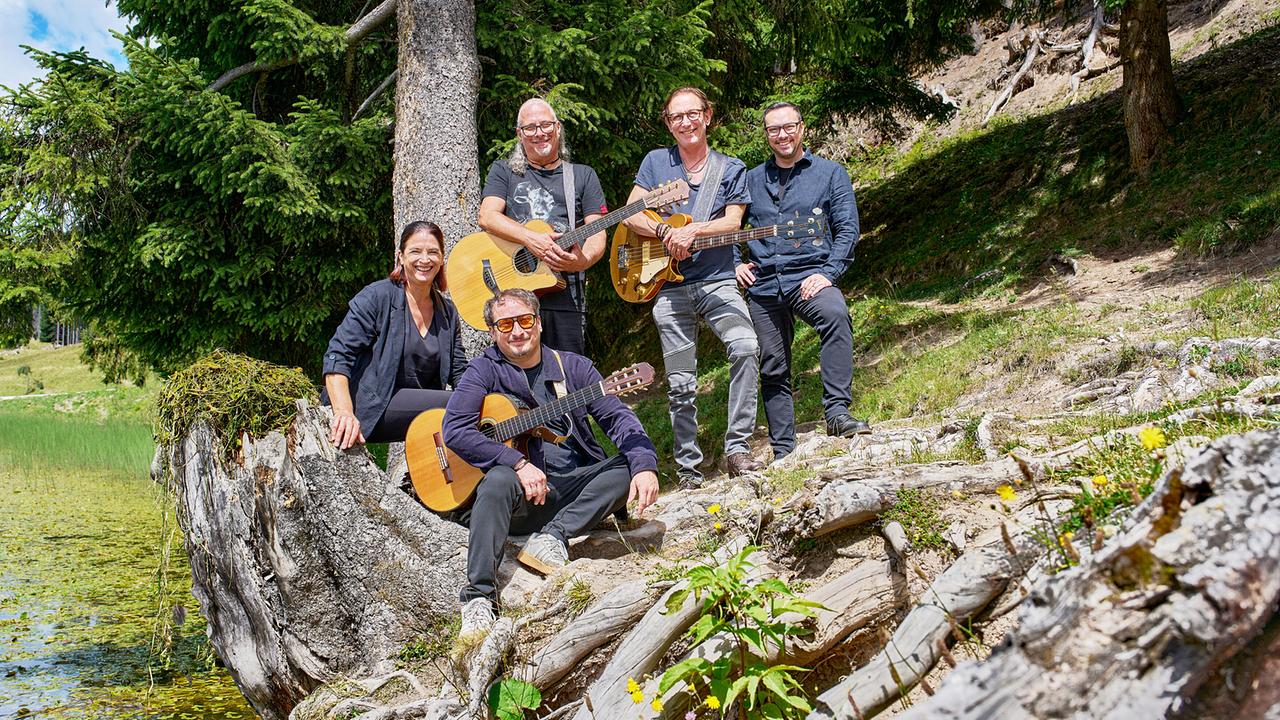 Krauthobel, fünf Musiker:innen mit Gitarren, posieren an einem idyllischen Bergsee zwischen Bäumen und Felsen.