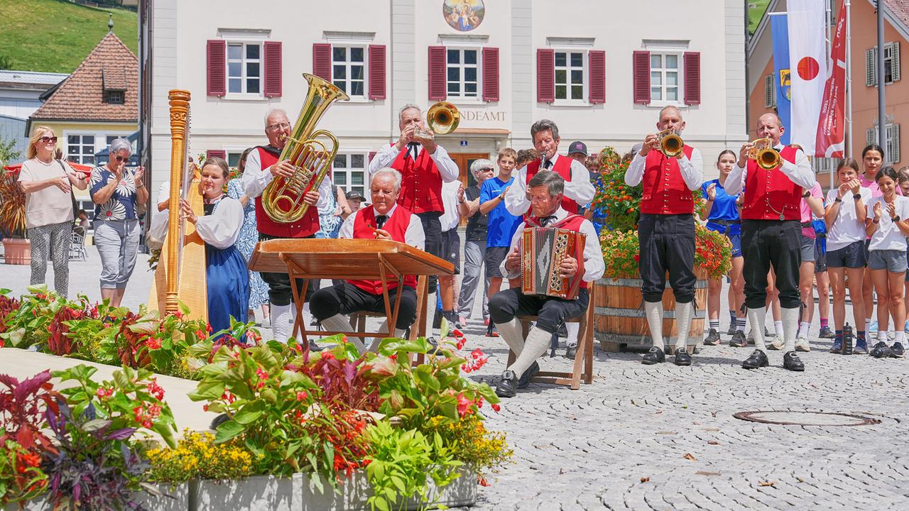 Die Muntafuner Tanzbodamusig, Musiker:innen in roten Westen, spielen vor einem Gemeindeamt auf einem festlich geschmückten Dorfplatz.