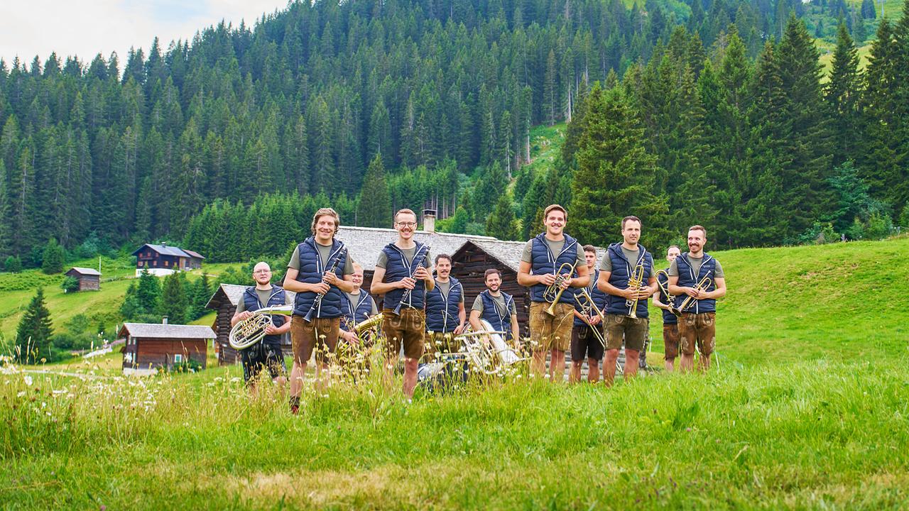 Die MuVukanten in Lederhosen steht mit ihren Instrumenten auf einer grünen Bergwiese vor bewaldeten Hängen.