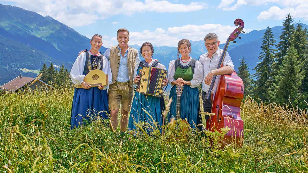 Hans Knauß posiert mit der Volksmusikgruppe Frosla Musig in Tracht und ihren Instrumenten vor einer alpinen Bergkulisse.