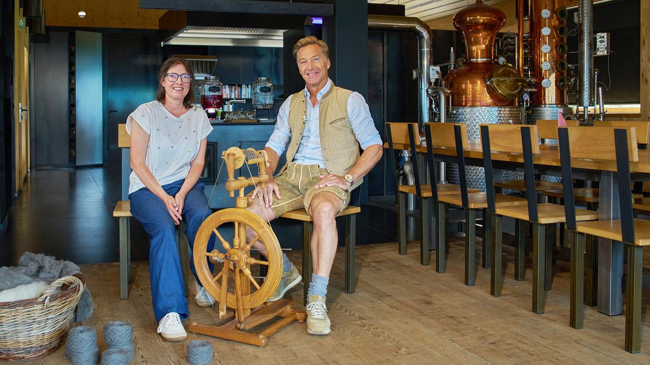 Doris Bitschnau und Hans Knauß sitzen neben einem Spinnrad in einer modernen Brennerei mit Kupferkessel.