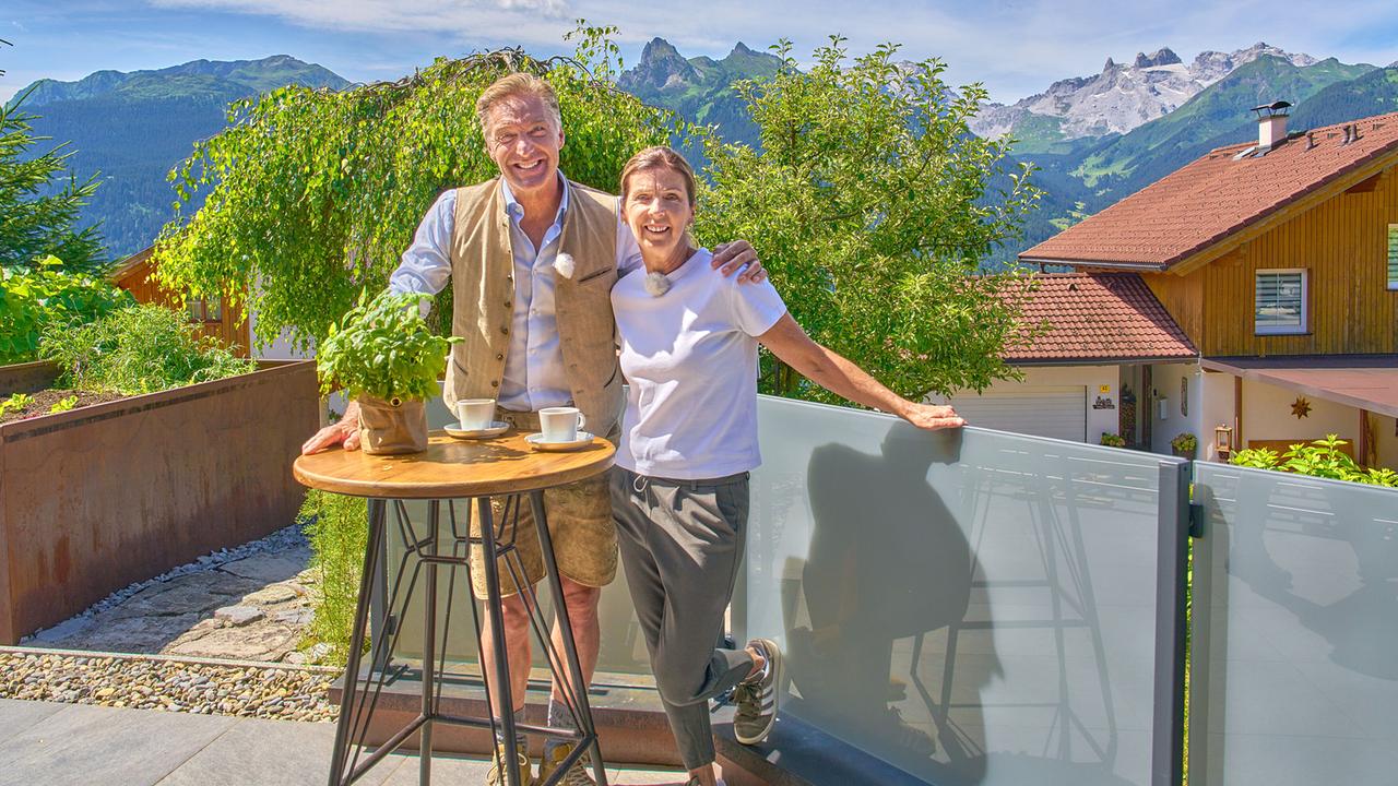 Hans Knauß und Anita Wachter stehen auf einer sonnigen Terrasse vor eindrucksvoller Bergkulisse.