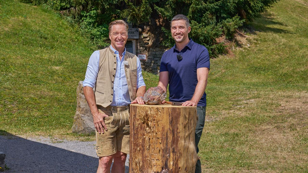 Hans Knauß und Christoph Walser  präsentieren im Freien ein Gesteinsstück auf einem großen Baumstumpf.