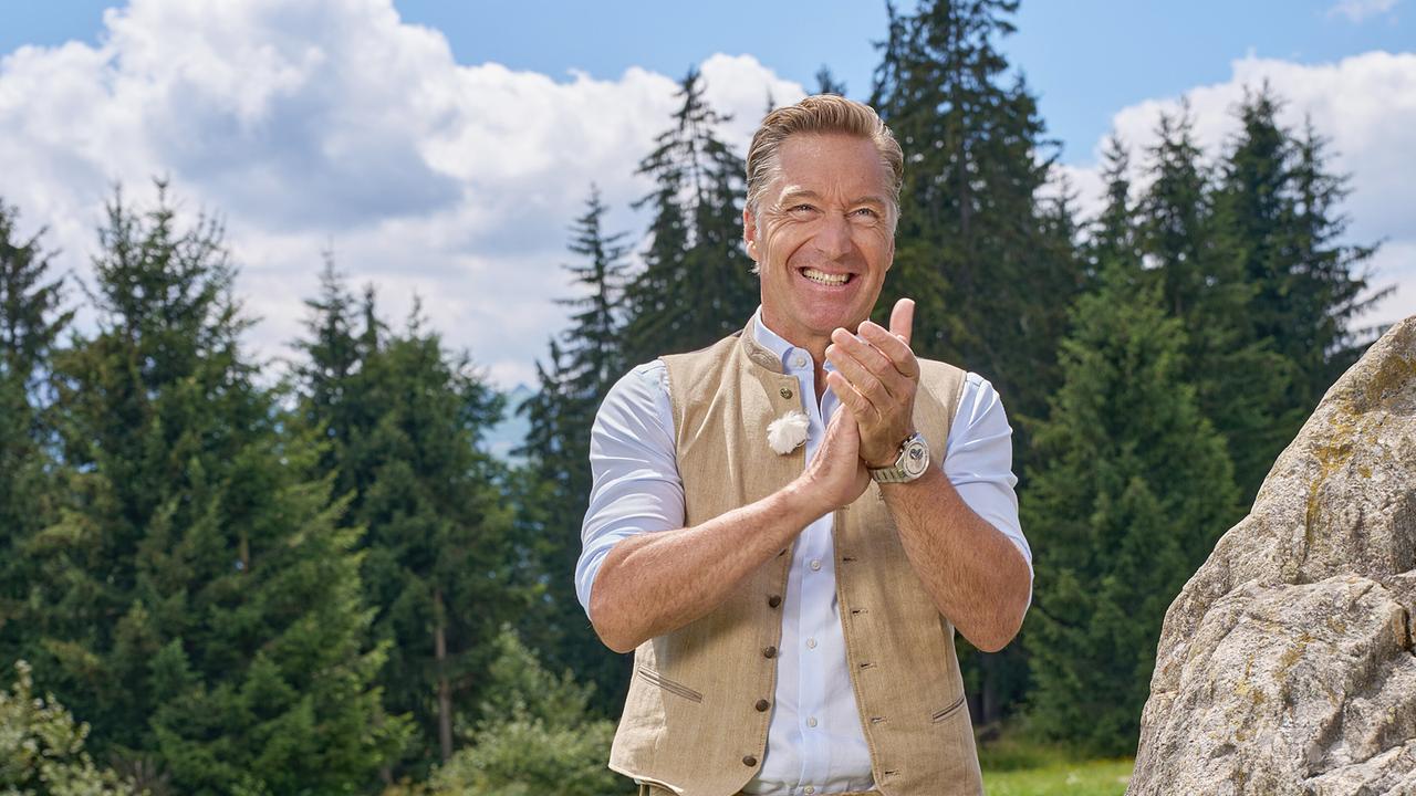 Hans Knauß klatscht lächelnd neben einem großen Felsen vor einem Wald.
