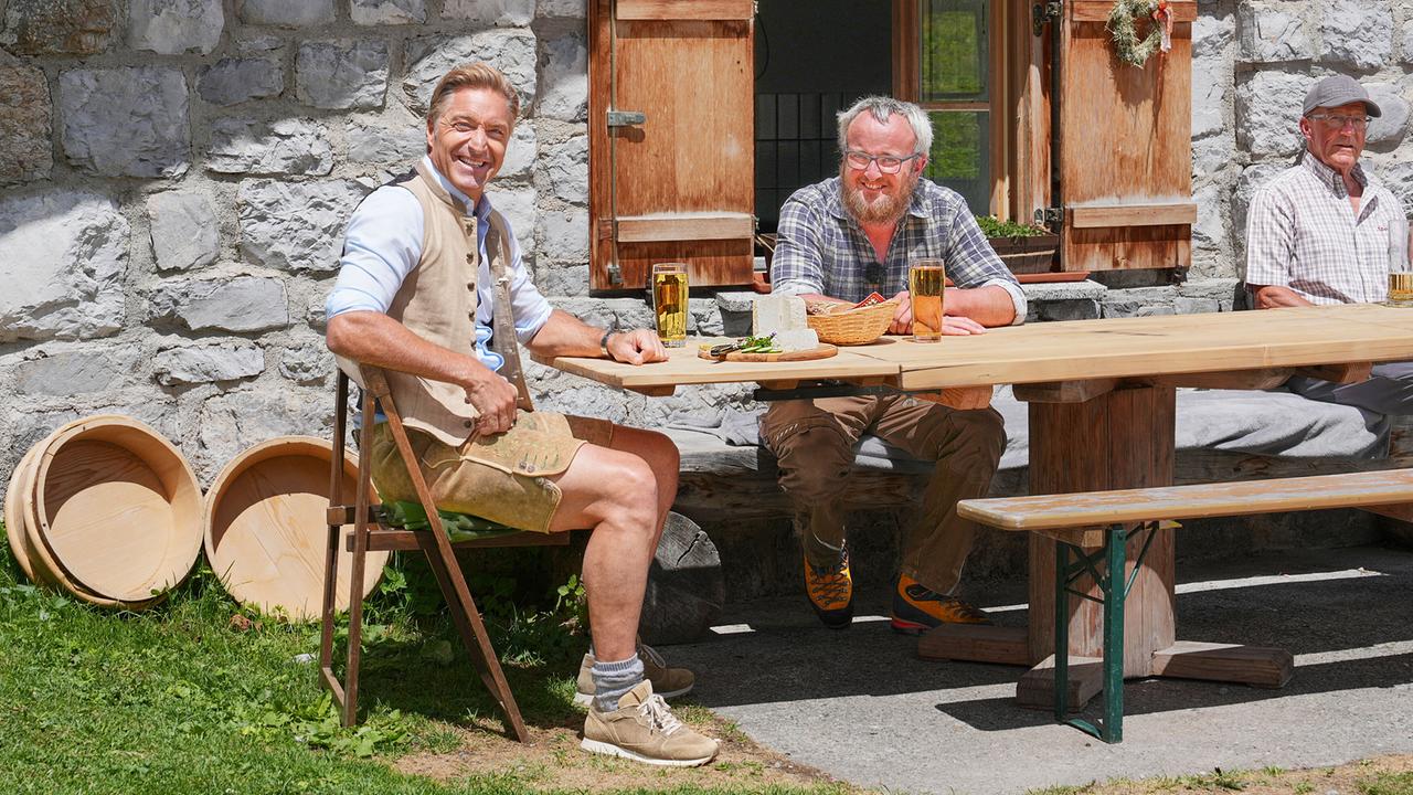 Hans Knauß sitzt mit Florian Battlogg vor einer urigen Steinhütte an einem Holztisch mit Getränken und Jause.
