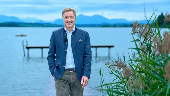 © ORF/Landesstudio Steiermark/Alois Kuschetz Hans Knauß steht lächelnd an einem Seeufer vor einem Steg, eingerahmt von Schilf und einer Bergkulisse.