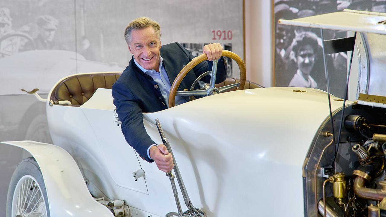 © ORF/Landesstudio Steiermark/Alois Kuschetz Hans Knauß sitzt lächelnd am Steuer eines historischen weißen Automobils in einem Museum.