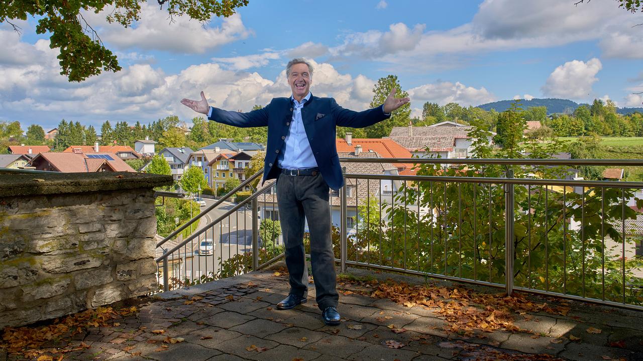 © ORF/Landesstudio Steiermark/Alois Kuschetz Hans Knauß steht mit ausgebreiteten Armen auf einer Terrasse über einem Ort in ländlicher Umgebung.