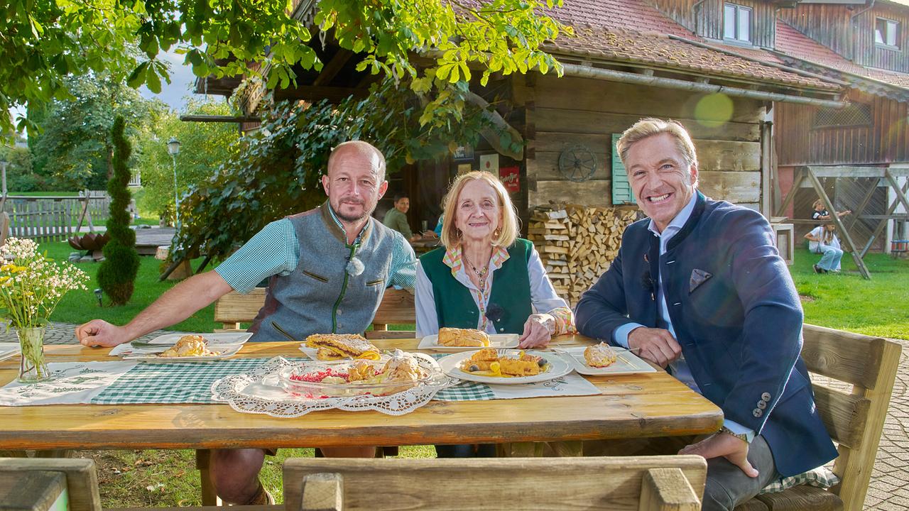 © ORF/Landesstudio Steiermark/Alois Kuschetz Hans Knauß sitzt mit Anton Greischberger und Hildegard Greischberger an einem Holztisch mit Mehlspeisen vor einem urigen Bauernhaus im spätsommerlichen Garten.