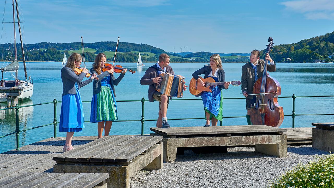 © ORF/Landesstudio Steiermark/Alois Kuschetz Die Seesaitn Musi, eine Volksmusikformation, spielt mit Geige, Harmonika, Gitarre und Kontrabass auf einem Steg am See.
