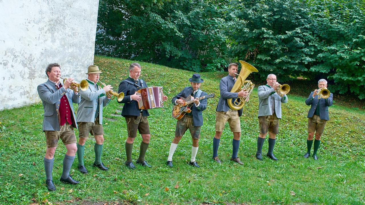 © ORF/Landesstudio Steiermark/Alois Kuschetz Flochgau Blech, eine Blasmusikgruppe in Tracht, musiziert auf einer Wiese neben einer alten Mauer.