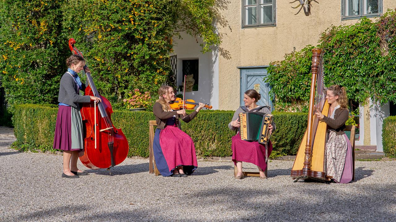 © ORF/Landesstudio Steiermark/Alois Kuschetz Die Mirabell Dirndln, vier Musikerinnen in Tracht, spielen mit Kontrabass, Geige, Harmonika und Harfe im Garten eines historischen Hauses.