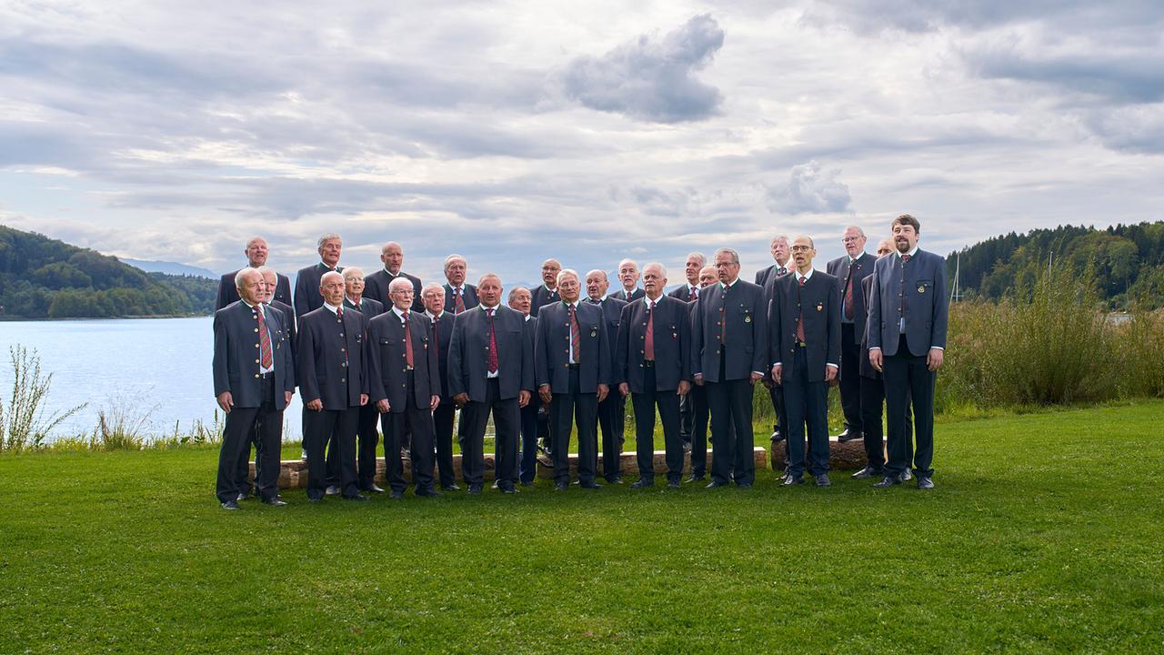 © ORF/Landesstudio Steiermark/Alois Kuschetz Die Liedertafel Neumarkt, ein Männerchor, steht in einheitlichen Trachtenjacken am Ufer eines Sees vor bewaldeten Hügeln.