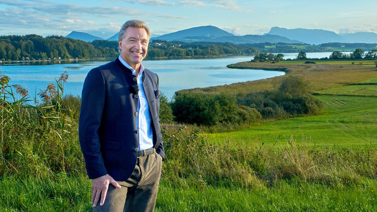 © ORF/Landesstudio Steiermark/Alois Kuschetz Hans Knauß steht lächelnd auf einer Wiese vor einem See und der Bergkulisse im Hintergrund.