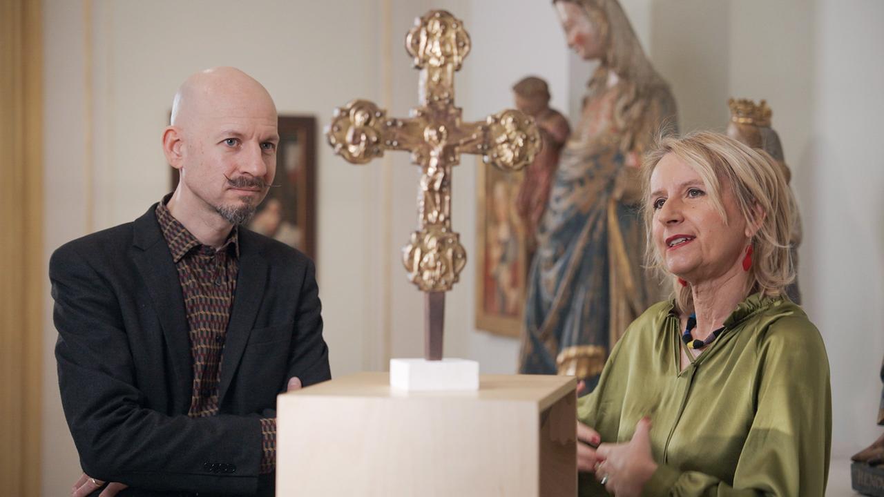 Filmemacher  Stefan Ludwig und Kunsthistorikerin Johanna Schwanberg stehen vor einem goldenen Kreuz im Dommuseum Wien