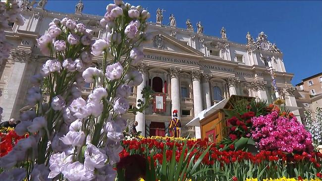© ORF Blumenarrangements schmücken den Petersplatz, während im Hintergrund der Petersdom mit einer aufgebauten Bühne zu sehen ist.