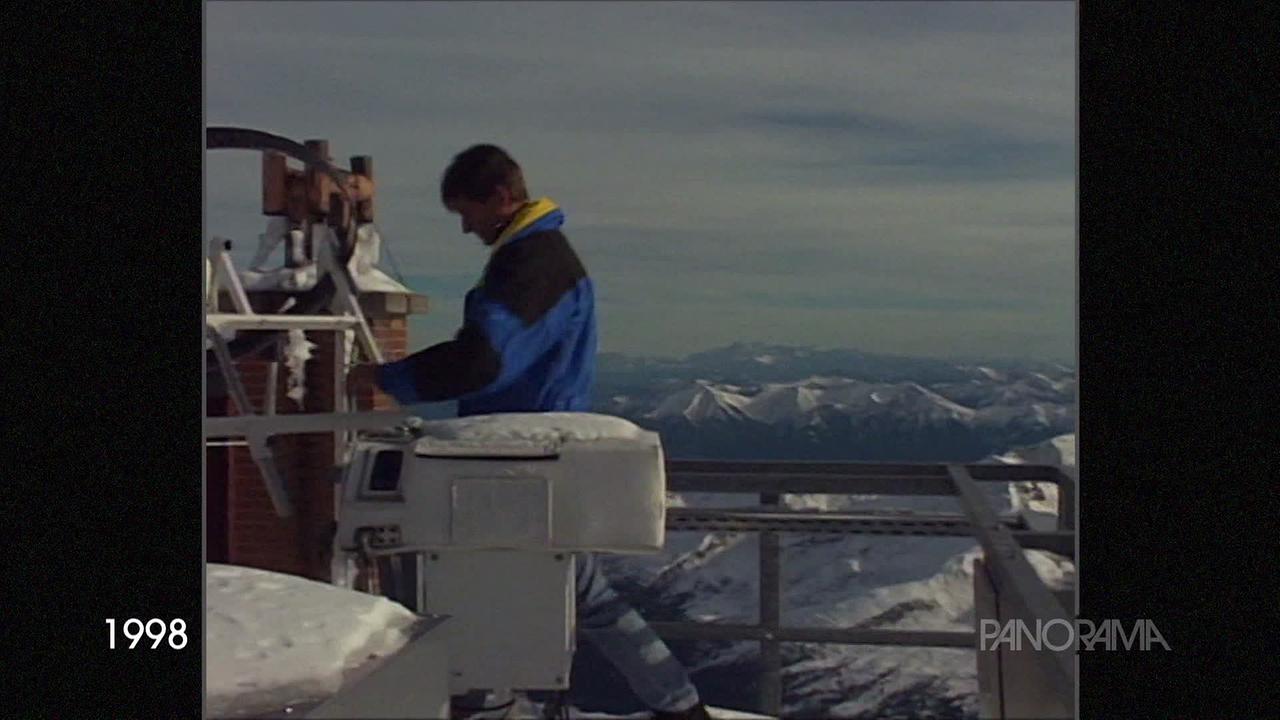 am Bild ist ein Mann im Anorak, der am Sonnblick Geräte im Außenbereich prüft. Es liegt Schnee und hinter ihm ist eine Aussicht auf viele schneebedeckte Gipfel.