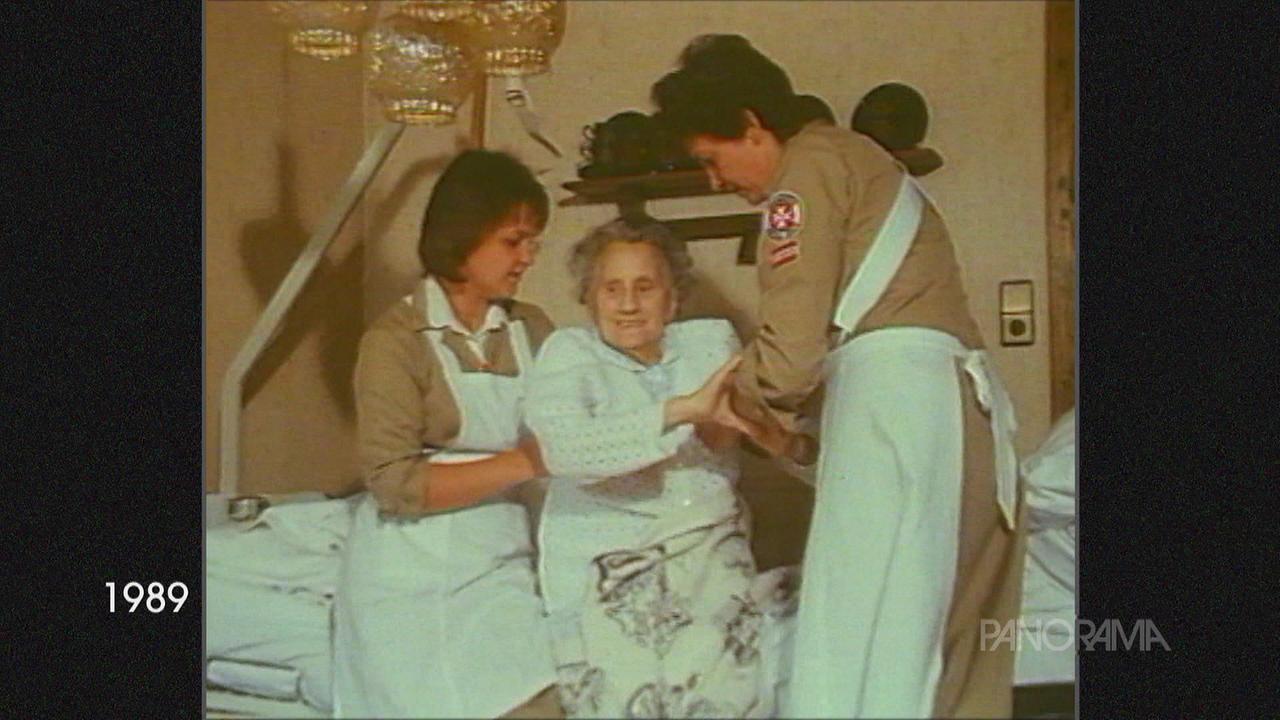 Zwei Helferinnen in Uniform und Schürze helfen einer alten Dame aus dem Bett.