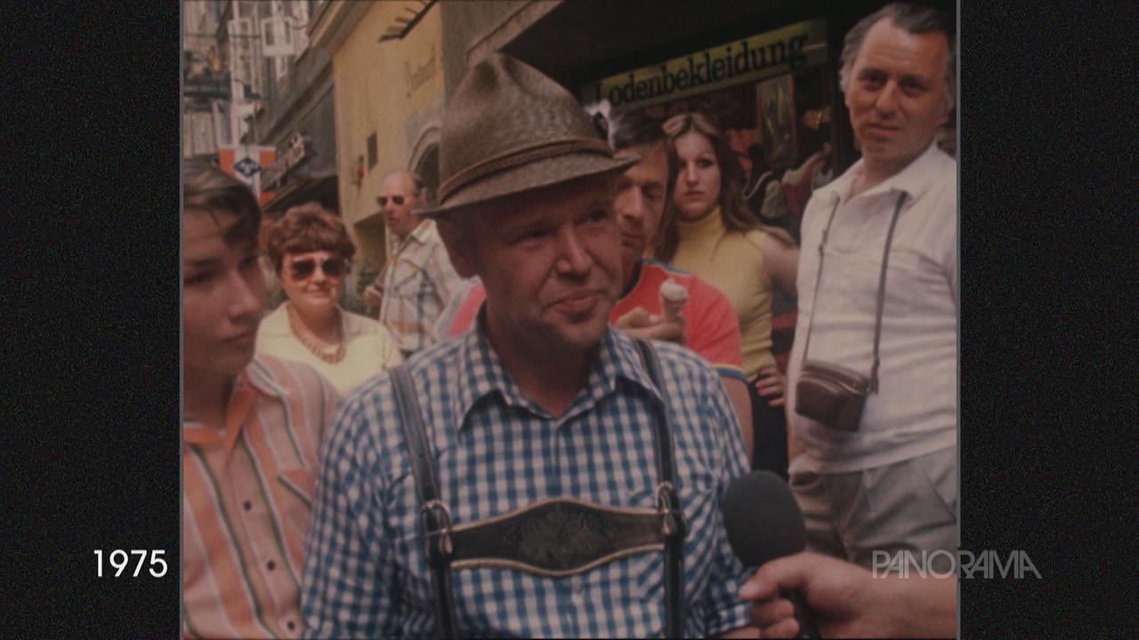 Am Bild von 1975 ist ein Mann mit kariertem Hemd, Lederhose und Hut im Interview zwischen vielen Menschen zu sehen. 