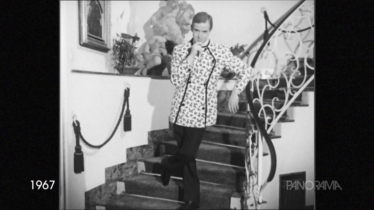 Am Schwarzweißfoto aus dem Jahr 1967 ist ein Herr in einer außergewöhnlich geschnittenen, langen Jacke auf einem Stiegenaufgang zu sehen. Die Jacke hat dunkle Ziernähte und Knöpfe und eine Art Leopardenmuster auf weißem Grund.