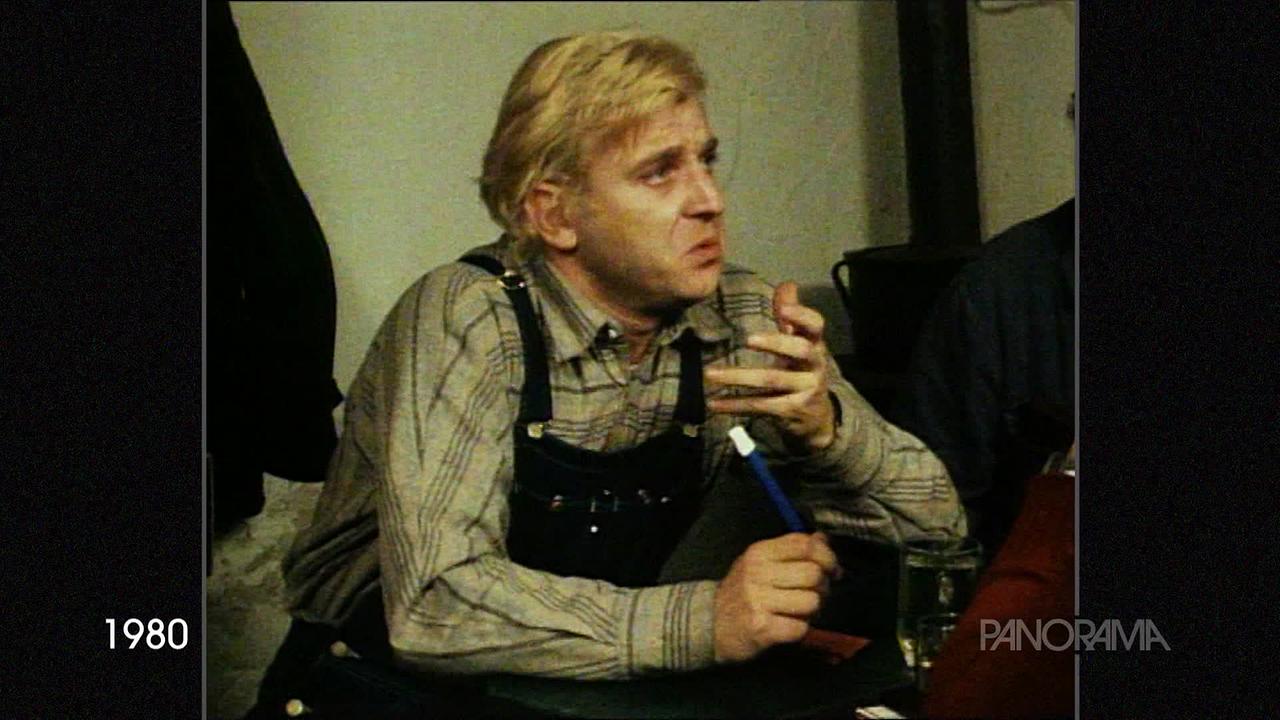 © ORF Am Bild aus dem Jahr 1980 ist Lukas Resetarits in einem karierten Hemd und Latzhose zu sehen. Er sitzt an einem Tisch.