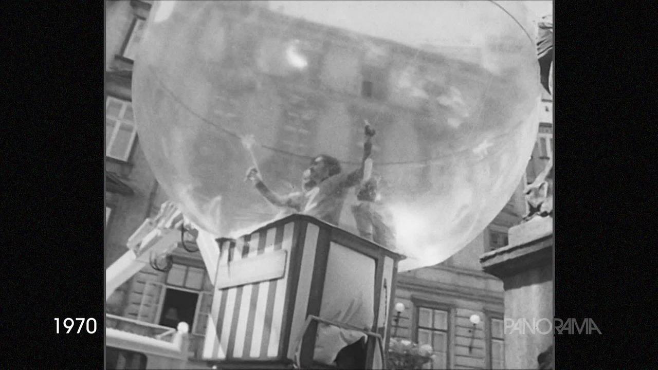 © ORF Am Bild aus dem Jahr 1970 ist ein durchsichtiger Riesenluftballon in dem Leute stehen, zu sehen. Drei Personen stehen auf einem Kranwagen in einem gestreiften "Ballonkorb" und im Ballon. Im Hintergrund sind Wohnhäuser zu sehen.
