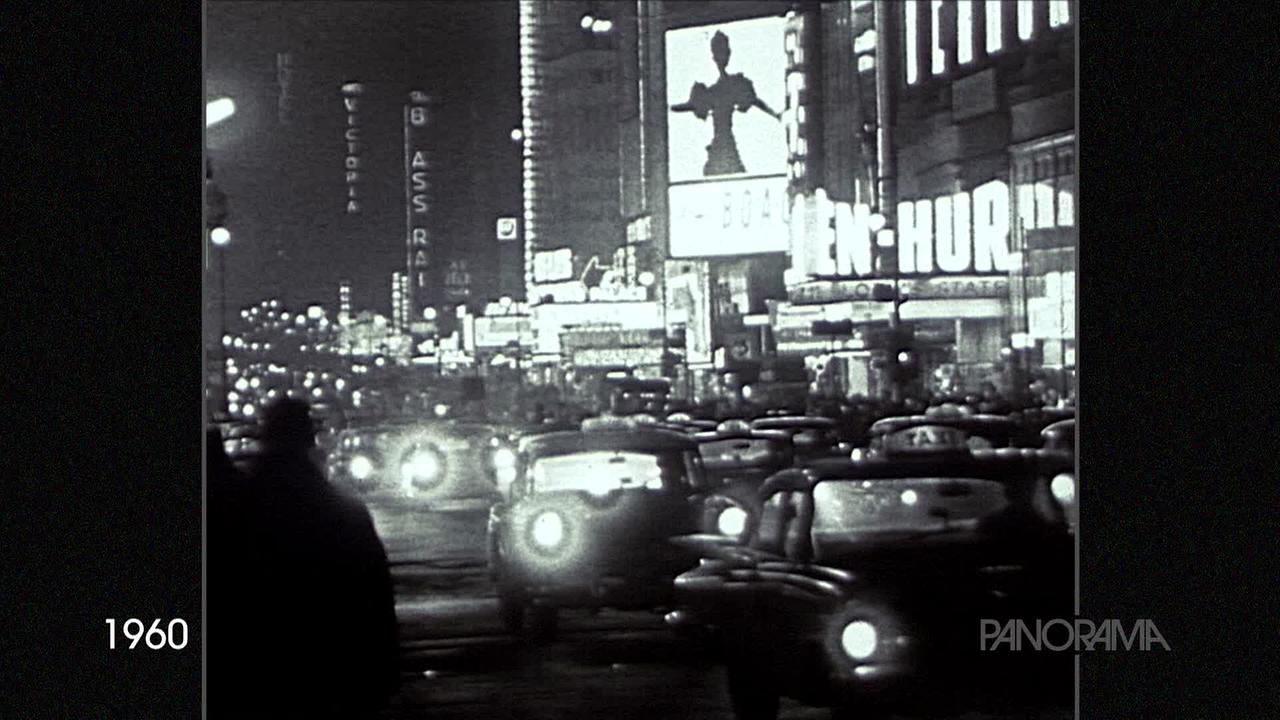 Am Schwarz-weiß-Bild aus dem Jahr 1960 ist eine dicht befahrene Straße mit einigen Taxis in New York zu sehen. Gleich rechts ist ein Kino in dem Ben Hur gespielt wird. im Hintergrund eine Menge Leuchtreklame.