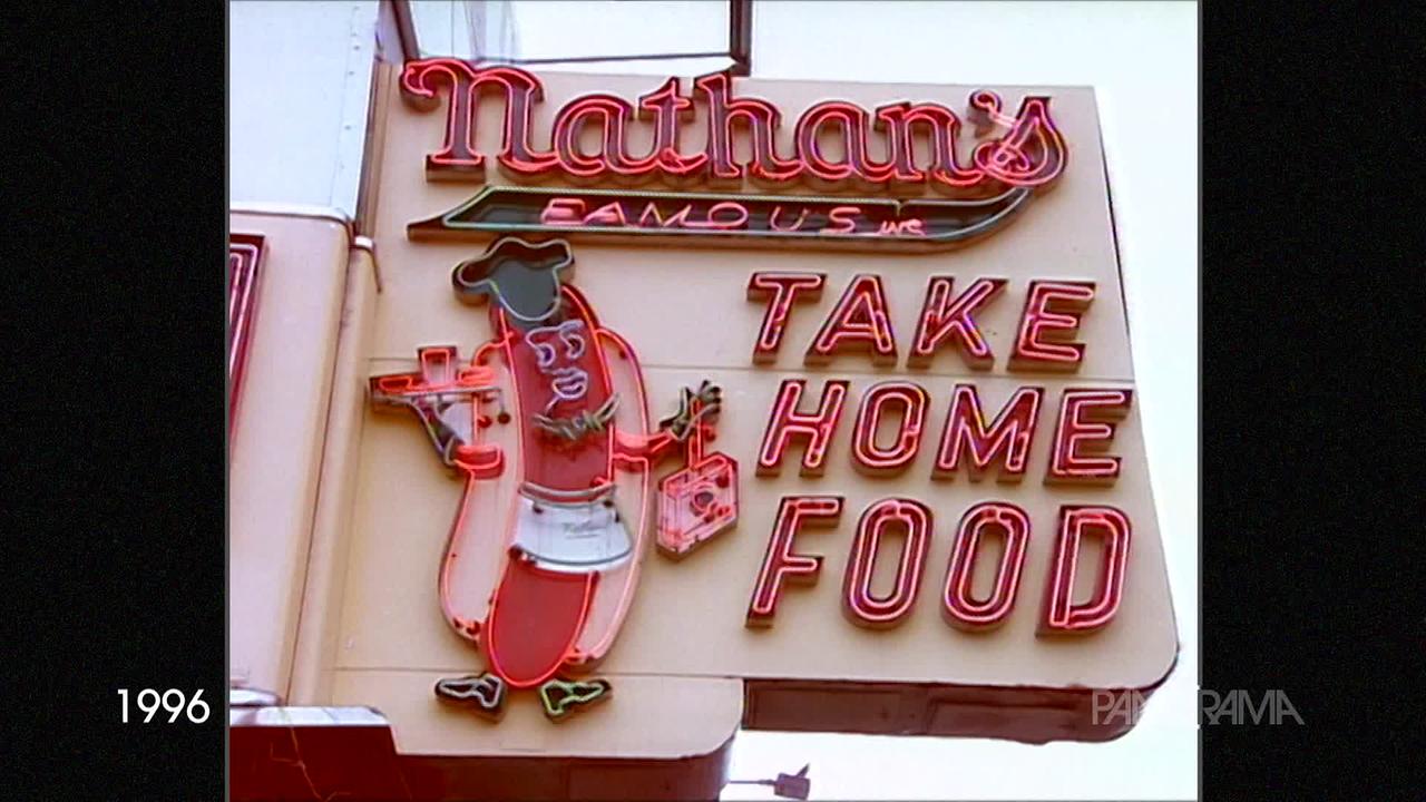 Am Bild aus dem Jahr 1996 ist eine Leuchtreklame von "Nathan's Take Home Food" zu sehen. Ein Hotdog mit Gesicht und Füßen trägt Essen und Getränke.