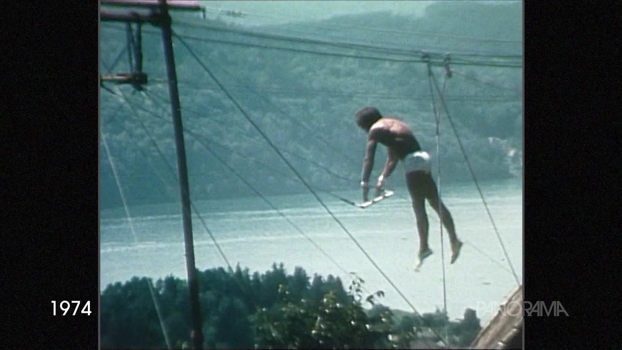 Am Bild ist ein Akrobat am Trapez im Freien zu sehen. Er trägt nur eine Short und schwingt hoch über den Baumwipfeln am Trapez. Im Hintergrund ist ein Gewässer zu sehen mit bewaldetem Ufer.