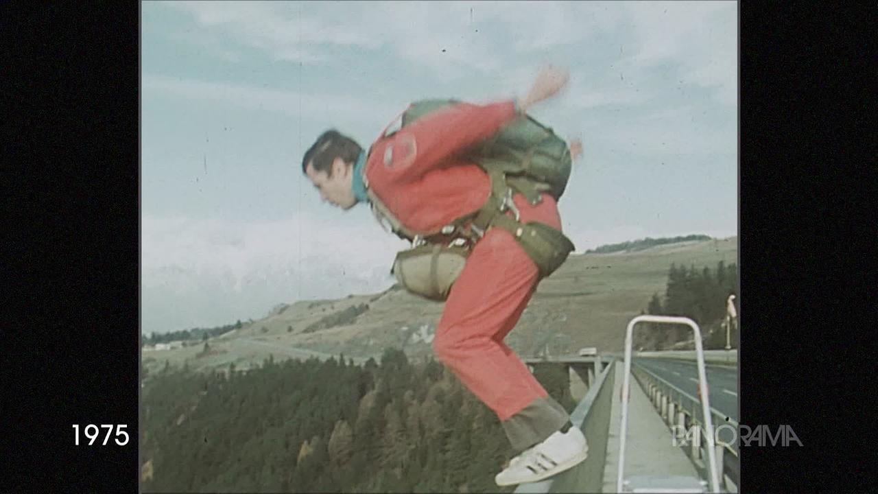 Am Bild aus dem Jahr 1975 ist ein Mann im roten Overall, der einen Fallschirm am Rücken hat. Er steht am Geländer einer Brücke und ist im Begriff zu springen.